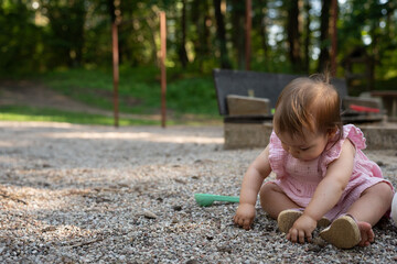 adorable infant in pink dress plays with pebbles on a playground surrounded by lush greenery under the dappled sunlight of a serene forest setting, wander, carefree, interaction, unstructured