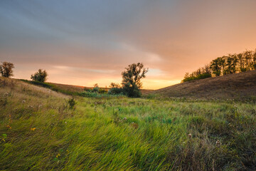 a picturesque landscape of rolling hills covered in a mix of lush green and golden grasses under a dramatic, multicolored sunset sky.
