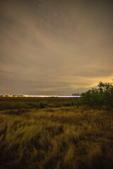 A long exposure of a night train passing through a field, creating a bright horizontal light trail, with faint stars visible through a cloudy sky.