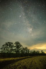 A long exposure of a night train passing through a field, creating a bright horizontal light trail, with faint stars visible through a cloudy sky.