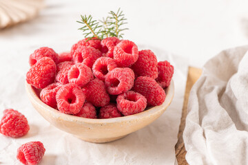 A rustic ceramic bowl filled with plump, fresh raspberries sits on a white table surrounded by scattered granola