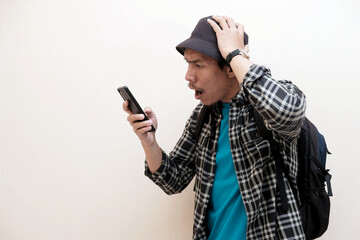 Southeast Asian man wearing cap and carrying backpack and looking to mobile phone that he hold with shocked expression, vacation, holiday.