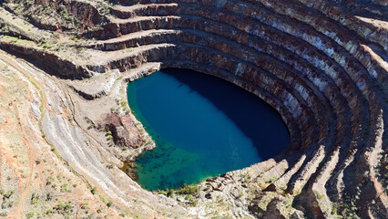 Aerial view of a deep blue crater lake of the Mary Kathleen Uranium Mine surrounded by steep rocky walls in a quarry. Turquoise water near the shore contrasts with the deep blue center