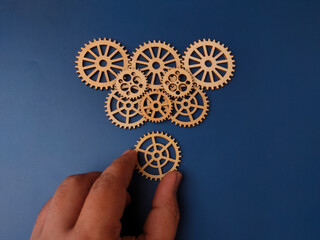 The image displays a hand placing a wooden gear among a collection of other wooden gears on a blue background