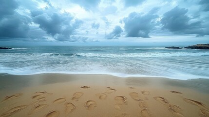 Footprints on sandy beach with gentle waves under a cloudy sky and rocky shoreline in the distance