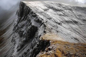 Rocky mountain ridge partially covered with light snow under foggy sky, showing sharp cliffs and earthy terrain