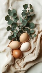 Four different colored eggs arranged on a soft beige fabric with eucalyptus leaves creating a natural, calm and organic atmosphere
