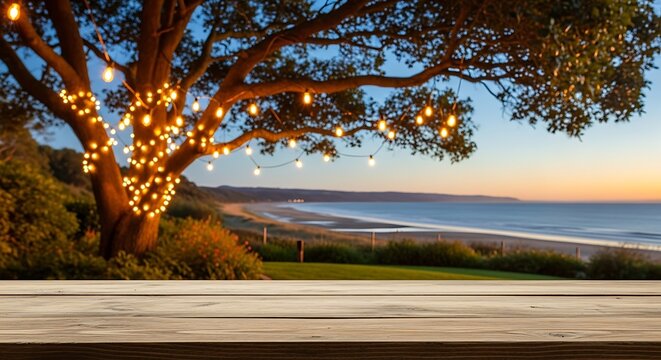 A wooden table top positioned in front of a scenic coastal view with a large tree adorned with warm string lights.