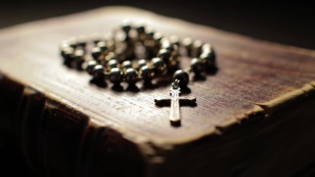 A close-up shot of a silver rosary with a cross resting on the worn cover of an old, leather-bound book, bathed in soft, dramatic lighting.