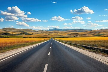 Fototapeta premium Long empty asphalt road stretching into golden fields and rolling hills under a bright blue sky with scattered white clouds, evoking calm and openness