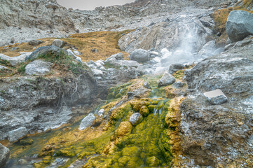Sulfur hot spring flowing from the Himalayas with green-yellow mineral markings on stones and rising steam, showcasing geothermal activity,Himalayan landscape, and natural wonder