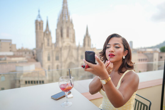 A woman is sitting at a table with a glass of wine and a cell phone. She is applying lipstick while looking at her reflection in a compact mirror. Concept of relaxation and self-care