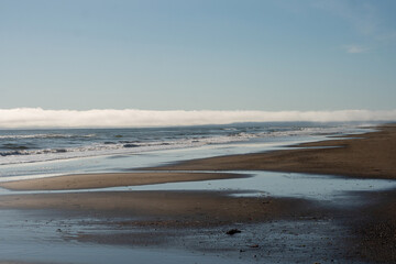 A sandy beach stretches into the distance with shallow pools reflecting the sky, while ocean waves meet a low fog bank along the horizon. Sea of Okhotsk, Sakhalin Island
