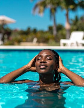 A woman floats peacefully in a turquoise pool, her hands resting on her head. The bright sunny day casts a warm glow, creating a serene outdoor atmosphere.