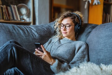 Obraz premium Young woman relaxing in cozy clothes enjoying music through headphones, lying on a comfortable sofa at home in a calm, peaceful mood.