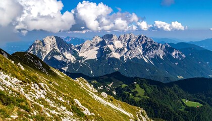 A vast vista unfolds, showcasing a mountain range with snow-capped peaks, lush valleys, and a vibrant green landscape beneath a partly cloudy sky.