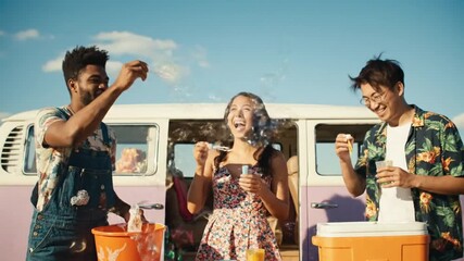Multi-ethnical group of hipster friends having a funny soap bubbles party close to a vintage van during a summertime road trip