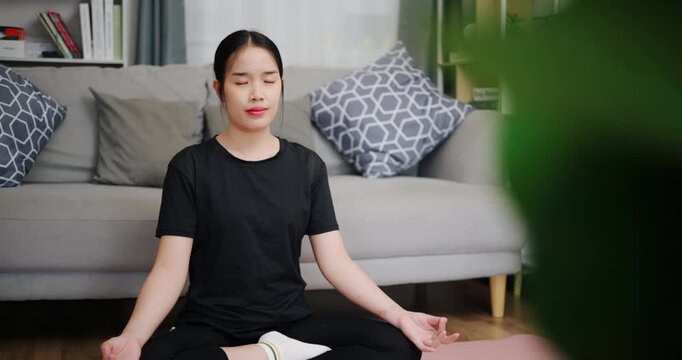 Handheld shot, Relaxed Asian woman practicing yoga by meditating on a yoga mat in living room at home. Self-care and mindfulness concept.