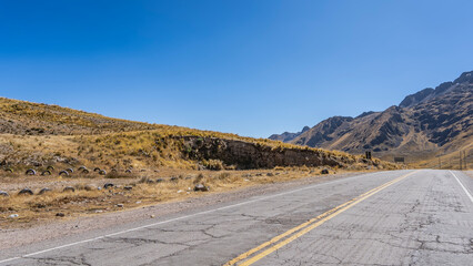 Highland highway. There is a double dividing line on the cracked asphalt. Dry, yellowed grass on the side of the road and on the hill. Mountains against a clear blue sky. Peru. The Andes Mountains.