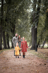 Fototapeta premium In an autumn park, two little girls are walking hand in hand on the road, while their parents stand embracing and watching them.