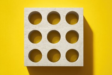 Off-white square block with nine evenly-spaced circular cutouts, casting a shadow on a vibrant yellow background.  The block appears to be made of wood or a similar material
