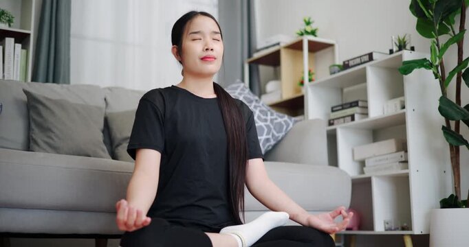 Handheld low angle view shot, Relaxed Asian woman practicing yoga by meditating on a yoga mat in living room at home. Self-care and mindfulness concept.