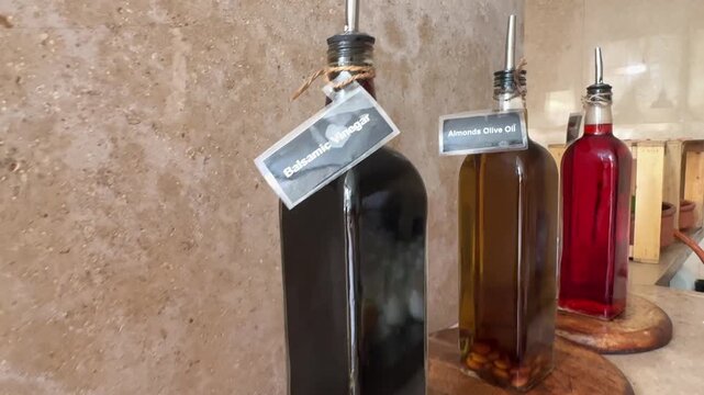 A variety of oil bottles displayed on a restaurant shelf. Labels show different types like olive, sesame, coconut, avocado, walnut, almond, flaxseed, grapeseed, and sunflower oils. Bottles are arrange