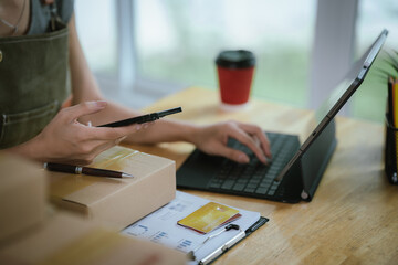 Fototapeta premium A young woman uses a credit card while working on a tablet at her home office desk, surrounded by parcels and stationery.