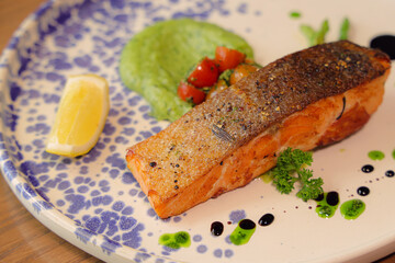 Exquisite Salmon Dish: A beautifully plated dish showcasing a perfectly cooked salmon fillet alongside vibrant green pea puree, fresh tomato, and a lemon wedge, presented on a patterned plate.