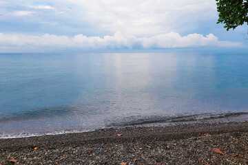 This photo shows a serene coastal view with calm sea water, a green hillside in the distance, and a small blue-roofed hut on the shore.