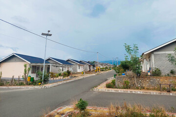 This image shows a row of small permanent houses in a housing complex, featuring blue and black metal roofs, small front yards, and colorful trash bins in front
