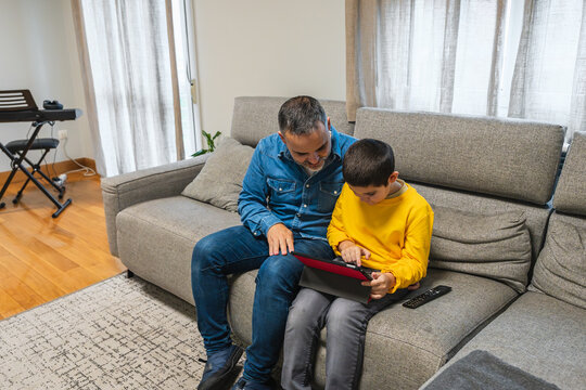 Father and son using a tablet together on a sofa in the living room, enjoying quality time and learning - Powered by Adobe