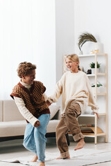 Two young children are dancing happily indoors in a bright living room, smiling and energetic, wearing cozy casual outfits amid soft natural light.