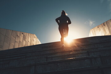 Low-angle shot of a woman running up long stadium stairs with sun flare behind her, dynamic composition symbolizing progress, perseverance, and determination in fitness and life.