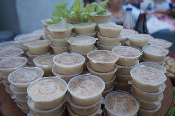 A large stack of Moustalevria desserts in plastic containers is shown at an outdoor event. This traditional cretan sweet pudding is infused with scented geranium (arbaroriza) and garnished with nuts.