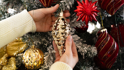 Close-up woman's hand holding Christmas gold ornament putting it on the tree, Christmas eve preparation  for winter holidays, front view