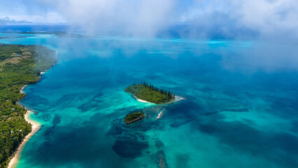 An atmospheric aerial view of the Isle of Pines lagoon in New Caledonia. A layer of mist or low cloud drifts over small, pine-covered islets sitting in the vibrant turquoise water © Anton Gots