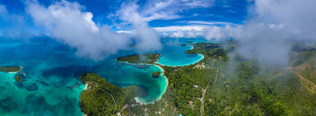 An expansive aerial panorama of the stunning Isle of Pines, New Caledonia. Clouds drift over the lush green landscape and the world-famous turquoise lagoons, revealing a tropical paradise