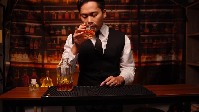 A professional bartender dressed in a stylish red vest and white shirt carefully holds a textured glass whiskey decanter and a glass of amber-colored spirit. The scene is set against a dark, moody bac