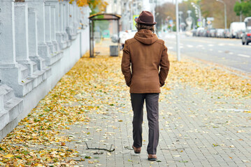 Man in brown coat and hat walking alone along city sidewalk covered with autumn leaves, seasonal change solitude urban lifestyle and peaceful outdoor atmosphere in fall