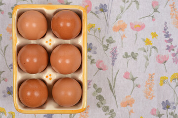 A close-up top-down view of a ceramic egg holder with six brown eggs, placed on a white tablecloth with a pastel floral print
