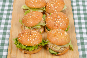 A group of six homemade burgers with sesame buns, lettuce, and cheese are arranged on a wooden board on a checkered tablecloth