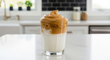 A glass of trendy whipped Dalgona coffee sits on a white marble countertop in a modern kitchen.