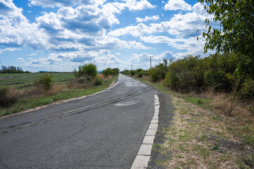 Curved rural road intersected by railway tracks, flanked by fields and power lines under a partly cloudy sky.