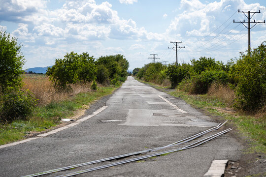 Curved rural road intersected by railway tracks, flanked by fields and power lines under a partly cloudy sky. - Powered by Adobe