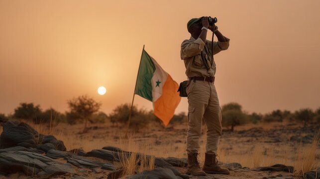 Focused male adult soldier using binoculars at sunset with flag in desert landscape military surveillance mission - Powered by Adobe