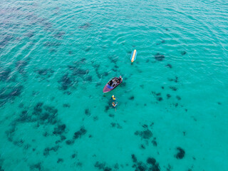 Scenic aerial view of a motorboat and a banana boat resting on calm blue-green waters while tourists with life vests enjoy swimming beside the boat.