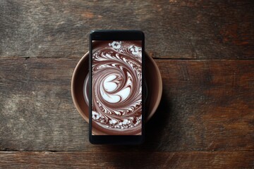 Artistic chocolate latte foam art displayed on a smartphone over a rustic wooden table background with a coffee saucer, blending technology and food art.