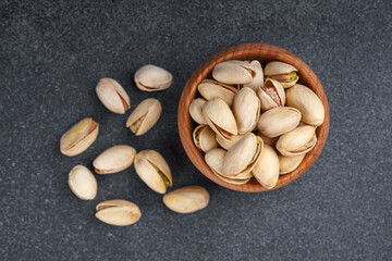 Close up, macro of salted pistachio nuts in wooden bowl with seeds scattered on textured dark gray background. Healthy snack food photography, organic nuts, natural texture, clean eating concept.
