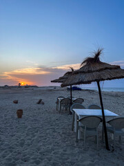 Enjoying a peaceful sunset over the blue waters of Djerba Beach in Tunisia, with thatched parasols providing shade for tables on the sandy shore.